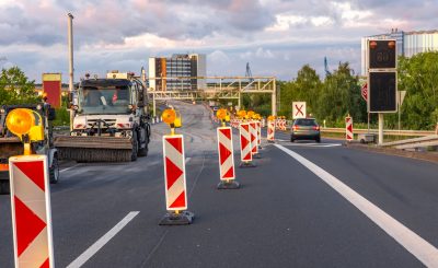 Road construction equipment and warning signs during highway repair against cloudy sky and modern architecture.