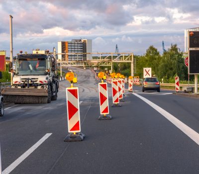 Road construction equipment and warning signs during highway repair against cloudy sky and modern architecture.