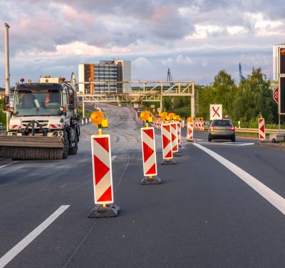 Road construction equipment and warning signs during highway repair against cloudy sky and modern architecture.