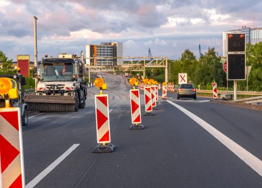 Road construction equipment and warning signs during highway repair against cloudy sky and modern architecture.